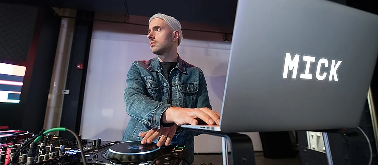 MICK behind a pair of Pioneer CDJs, hand on the jog wheel, a laptop lid reading MICK beside him
